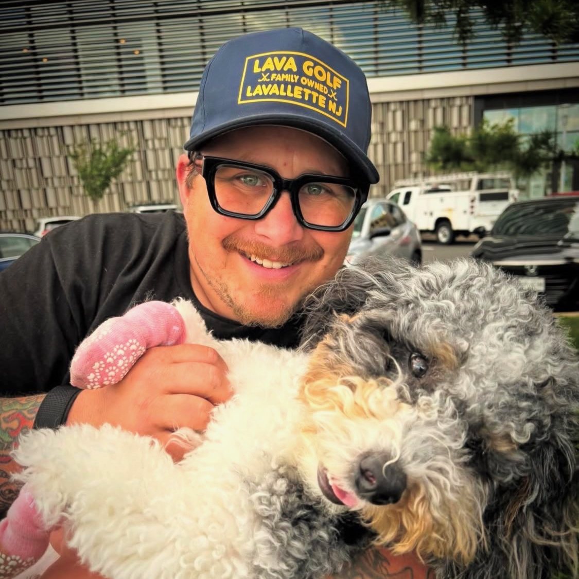 Chaz smiling in a ball cap and glasses, holding a merle doodle whose front paws are wrapped in pink bandages