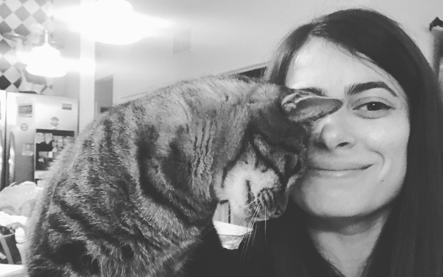 Black-and-white photo of a tabby cat pressing its head against a smiling woman's cheek in a kitchen